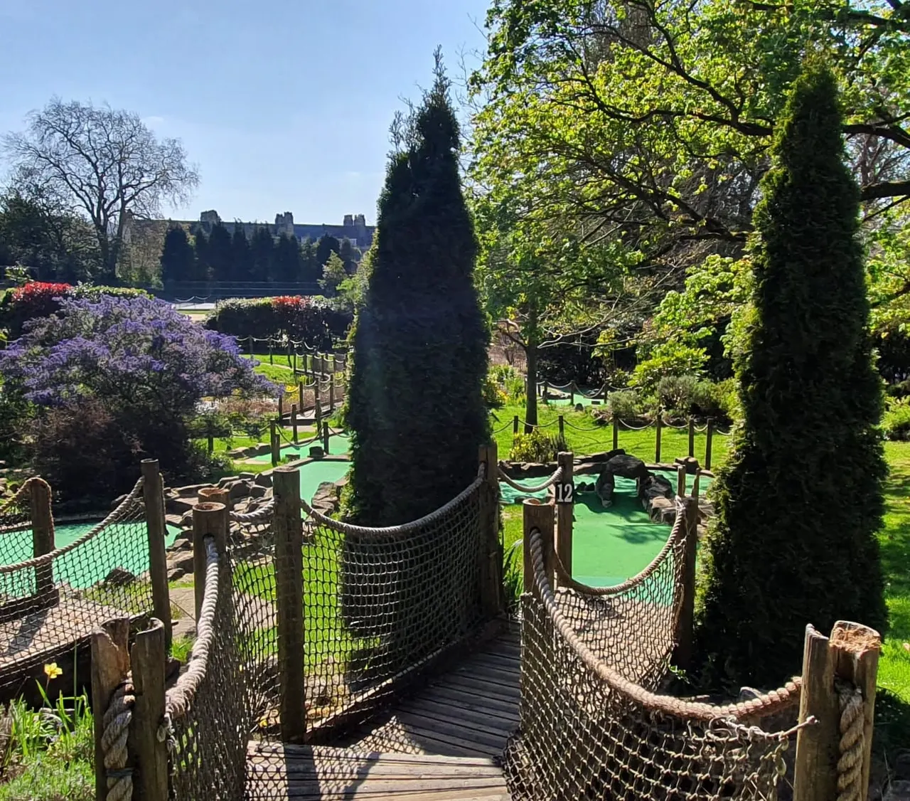 Rope bridge pathway with green mini golf course, tall dark hedges, flowering shrubs and castle visible in distance.