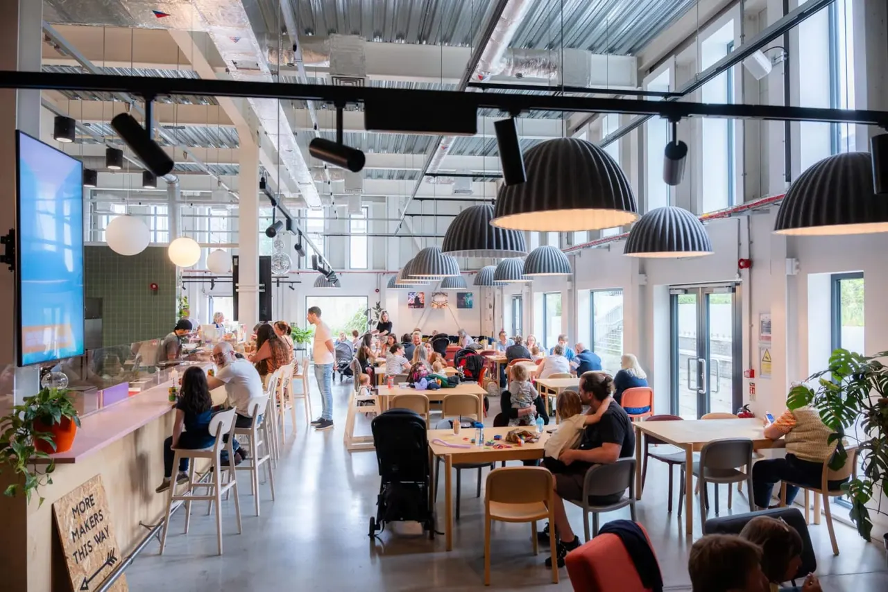 Busy café interior with mixed-age visitors dining and socialising at wooden tables beneath black dome pendant lights in a bright, modern space.