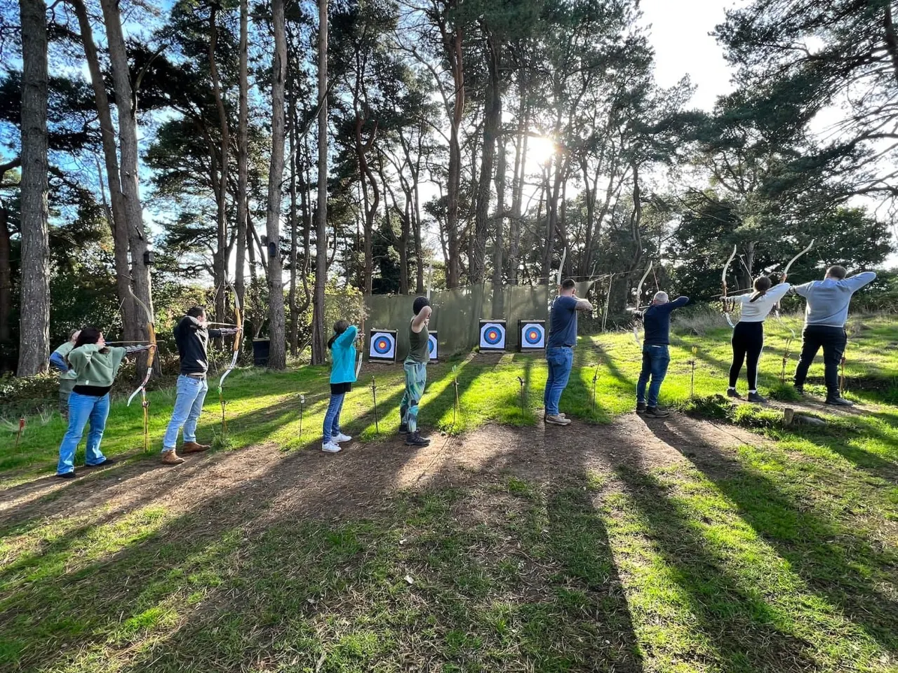 Group of people practising archery outdoors among tall trees, aiming at target boards in a woodland clearing.