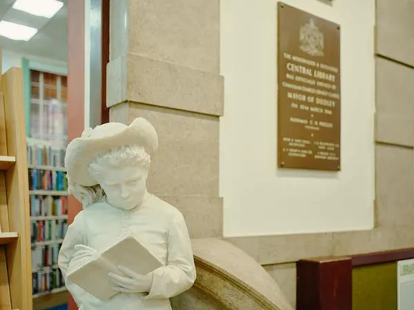 White marble statue of a child reading a book, displayed in a library interior with bookshelves and a commemorative plaque visible.