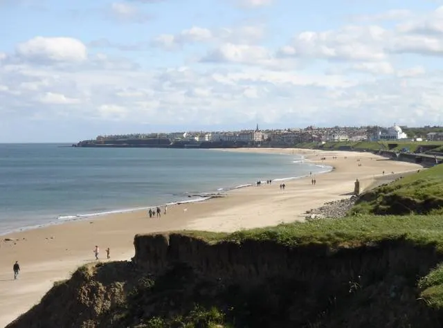 Sandy beach with scattered visitors, rocky cliffs in foreground, coastal town and buildings visible across the bay under partly cloudy sky.