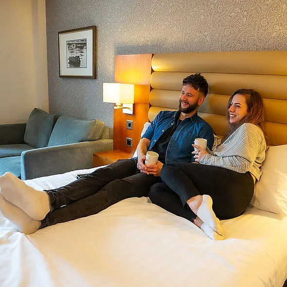 A man and woman sit together on a hotel bed, holding drinks and smiling at each other in a modern bedroom with gold headboard.