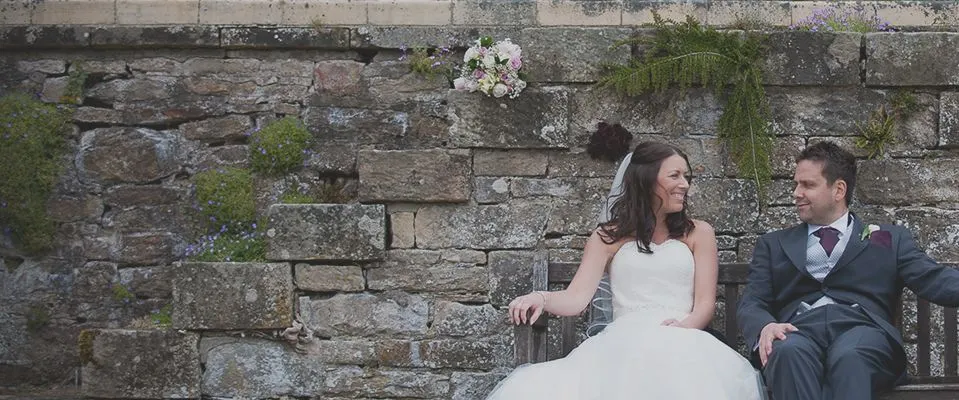 Bride and groom sitting together on a bench against a moss-covered stone wall with white flowers above.
