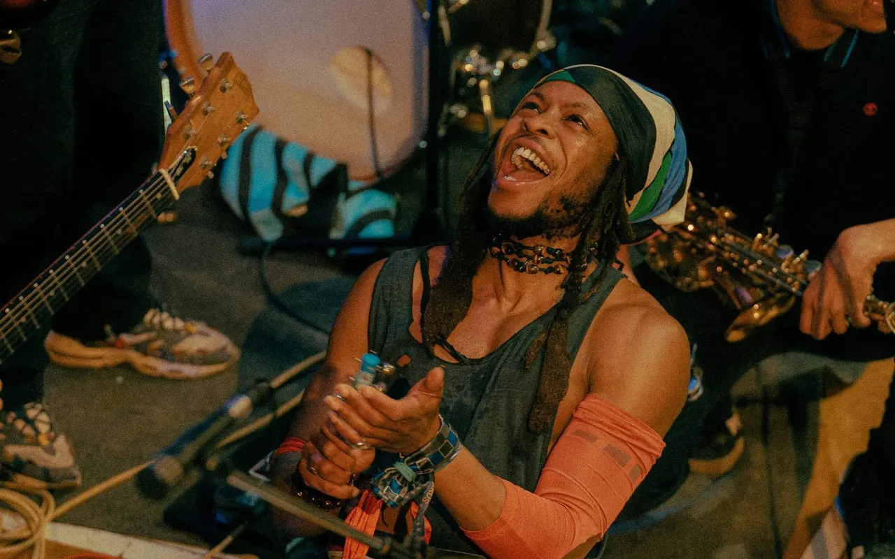 Musician performing with guitar and microphone on stage, wearing a colourful cap and studded accessories, smiling whilst singing.