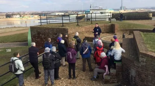 A group of visitors gathered around a guide during an outdoor tour of a historic fort, with fields and distant buildings visible beyond.