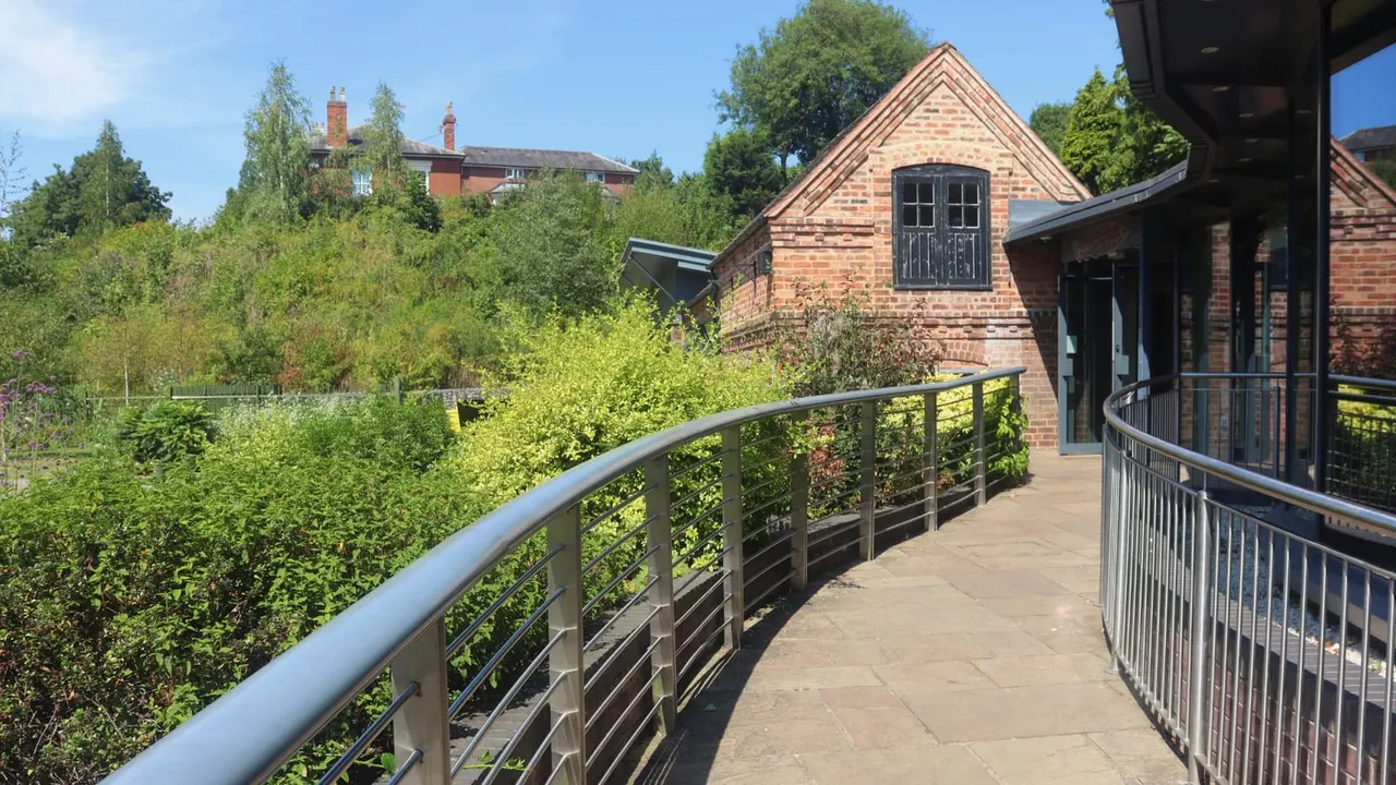 Paved walkway with metal railings curving past brick buildings surrounded by lush green trees and vegetation.