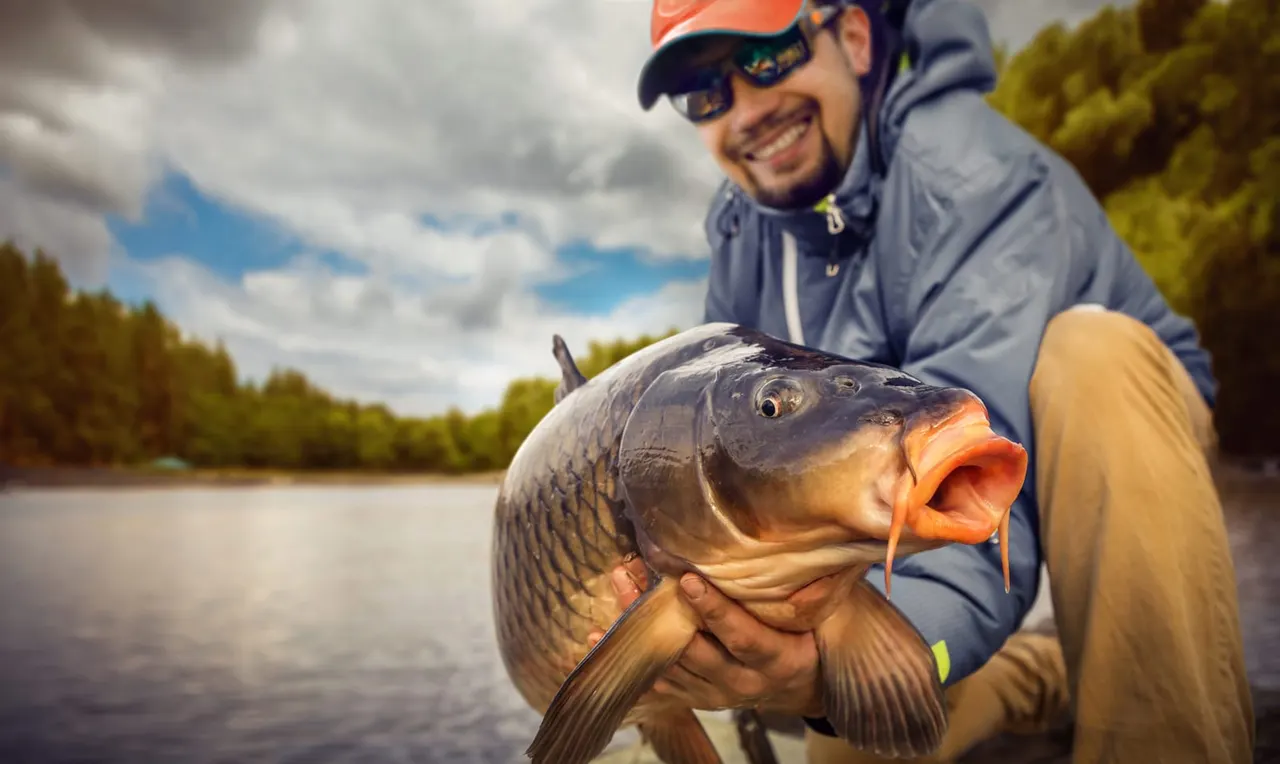 A man in a cap and sunglasses holds up a large carp he has caught whilst fishing by a river.