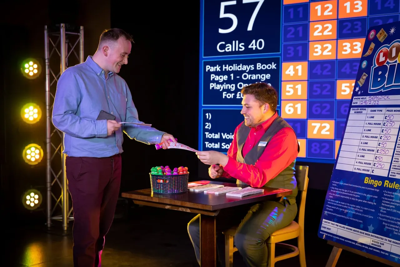 Two men playing bingo at a table with a large illuminated display board showing numbers and prizes behind them.