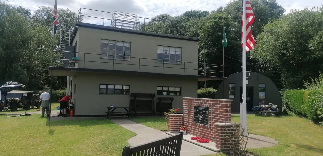 A modern museum building with distinctive architecture set in green grounds, featuring flags and a brick memorial with red poppies.