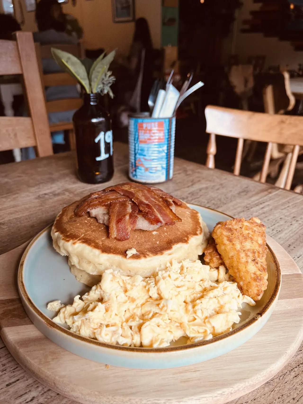 Plate with bacon and cheese toastie, scrambled eggs, hash brown, beer bottle and straws on restaurant table.