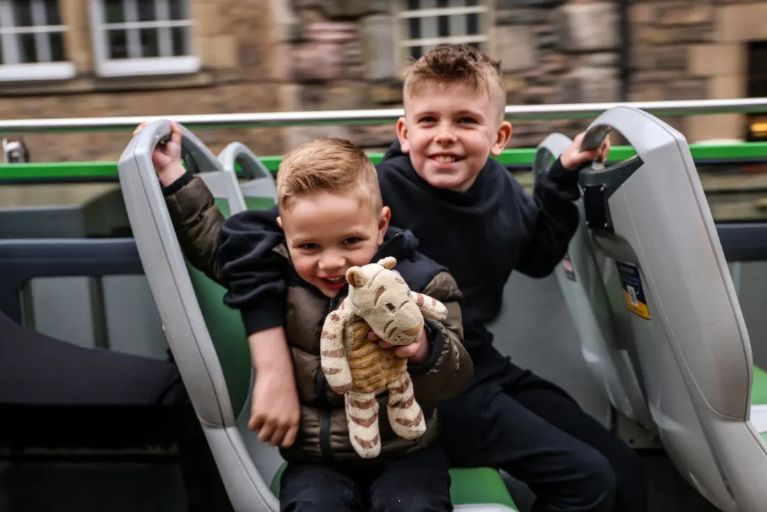 Two young children on a open-top bus ride, one holding a stuffed animal toy, smiling at the camera.