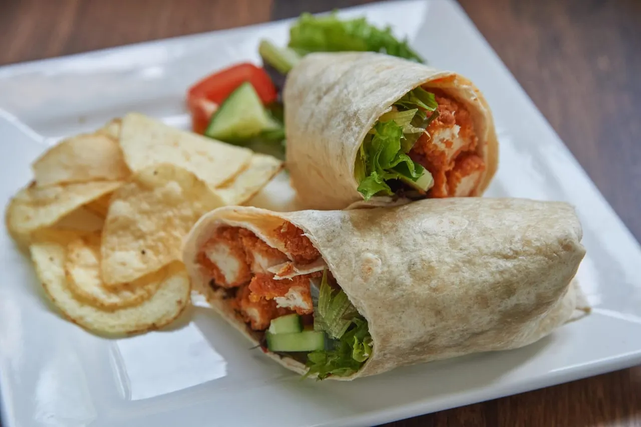 Two wraps filled with lettuce and tomato served with crisps and fresh vegetables on a white plate.