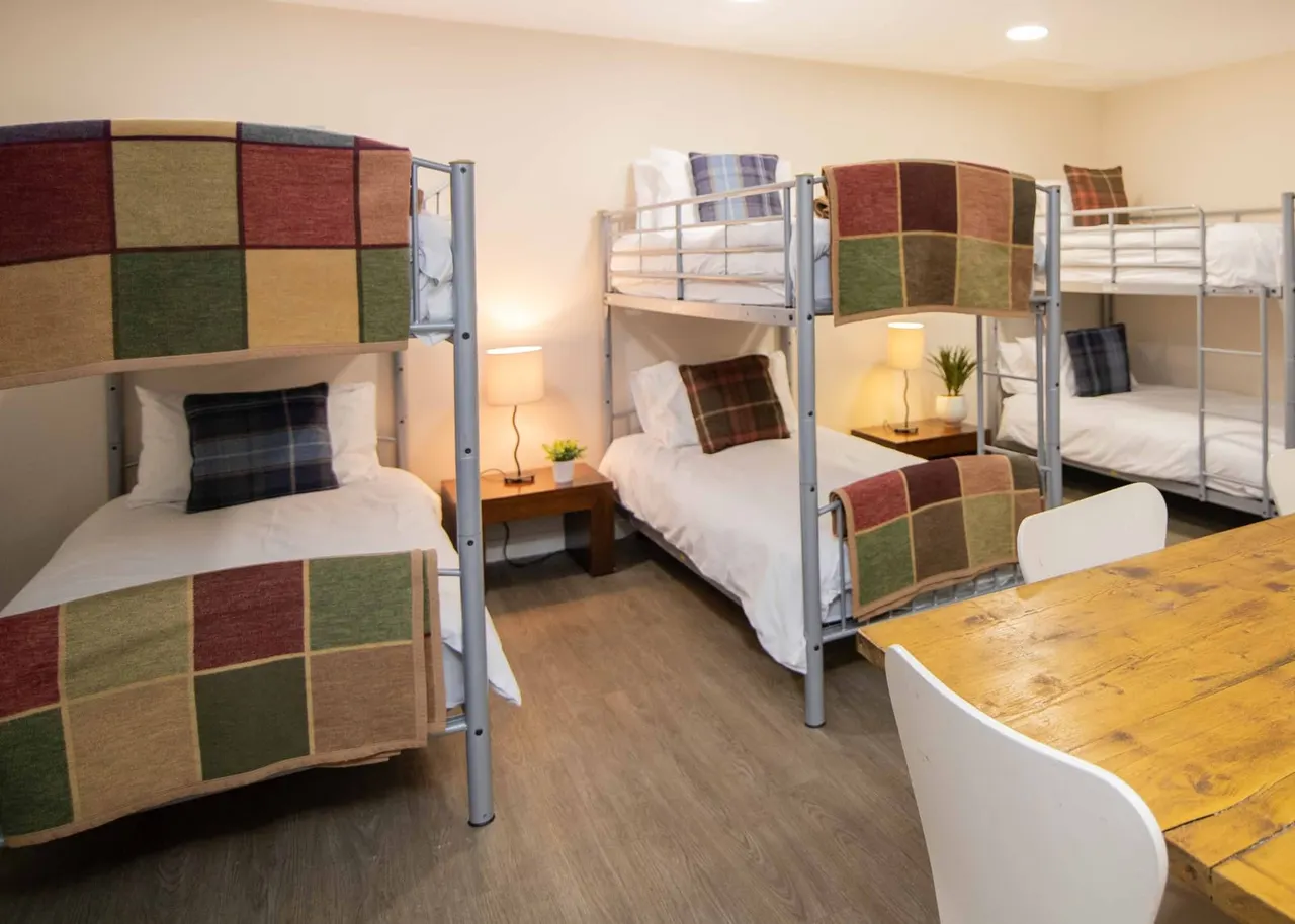A spacious dormitory room with multiple bunk beds with colourful tartan blankets, wooden table, and warm lighting.