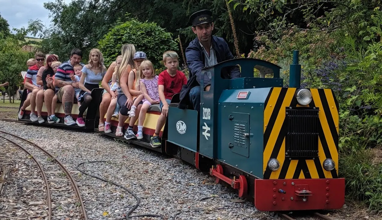 Families and children riding on a miniature railway train driven by a staff member through a leafy garden setting.