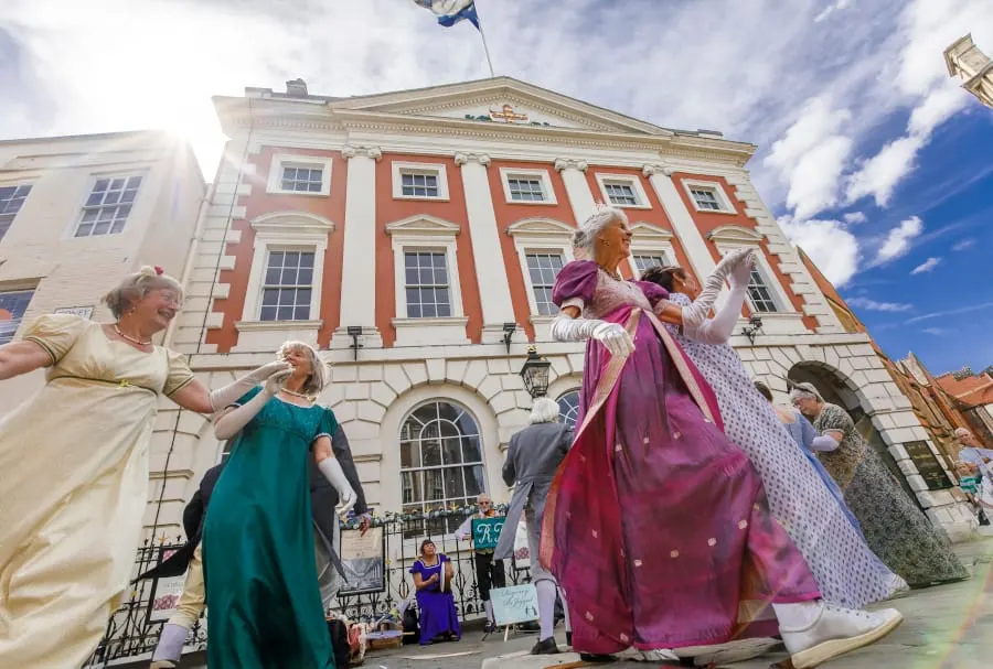 Costumed performers in period dress dancing in front of a Georgian red-brick mansion house building on a sunny day.