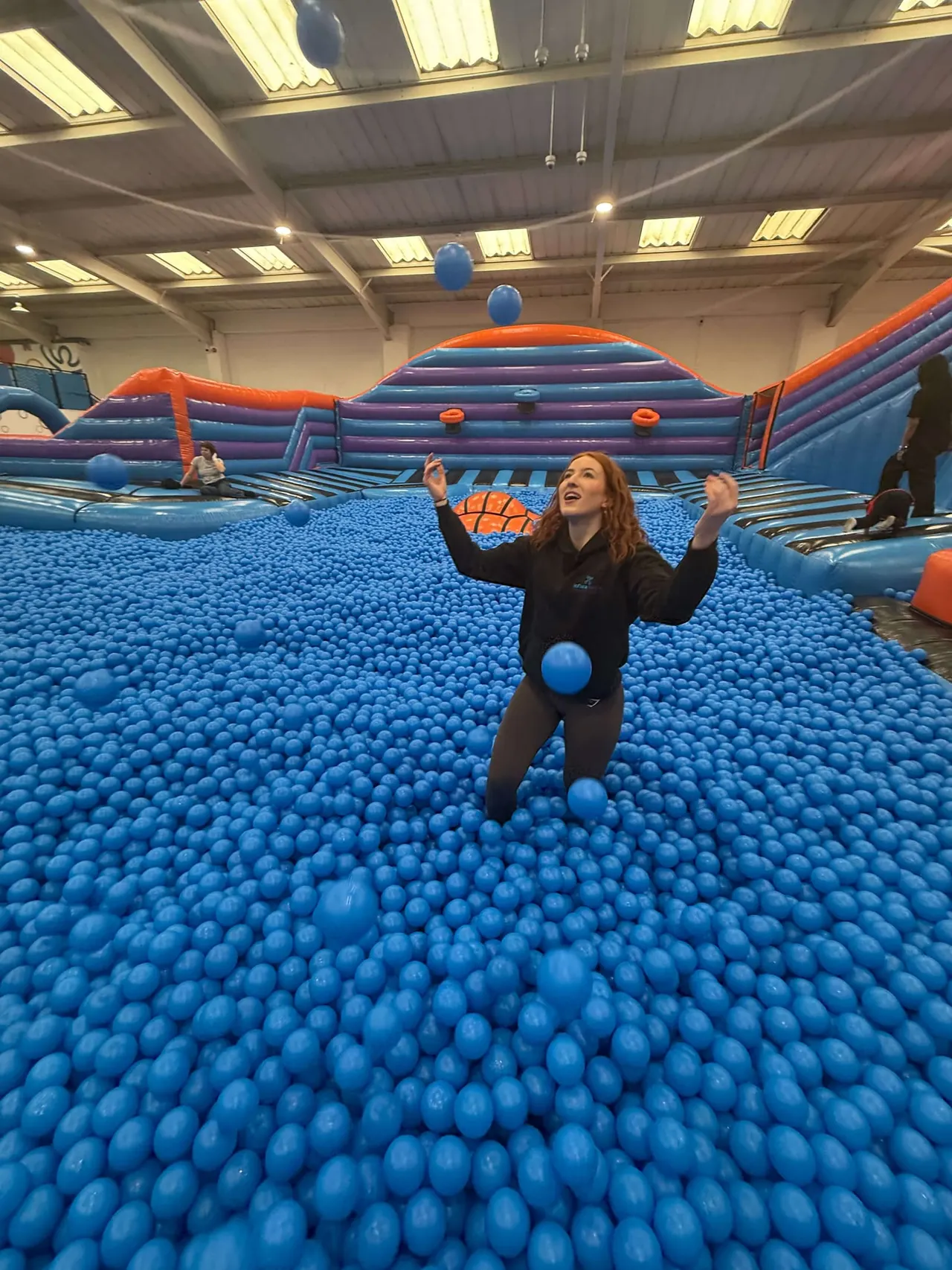 Woman playing in a vast ball pit filled with thousands of blue balls at an indoor trampoline park.