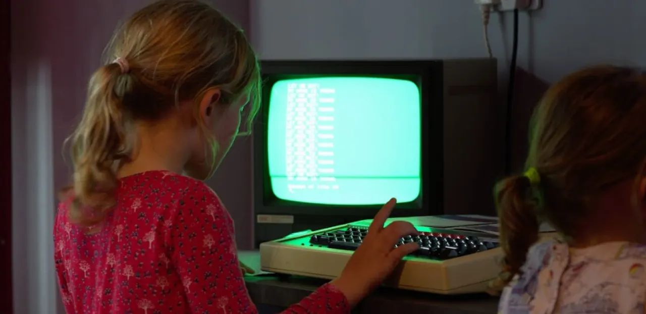 Two children using a vintage computer terminal with a green monitor displaying retro game graphics.