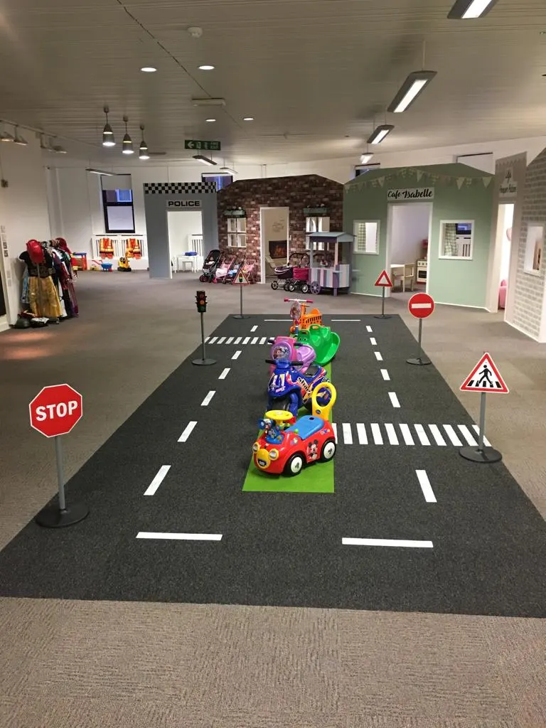 Indoor play area with toy vehicles on a road-marked floor and themed activity stations including a police station and café.