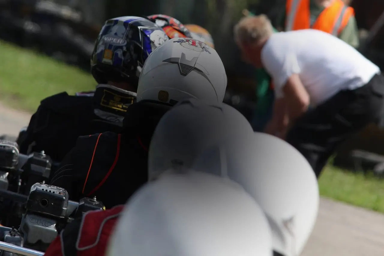 Go-kart racers wearing helmets line up with their karts on a grass circuit, with an official in orange high-visibility clothing overseeing the event.