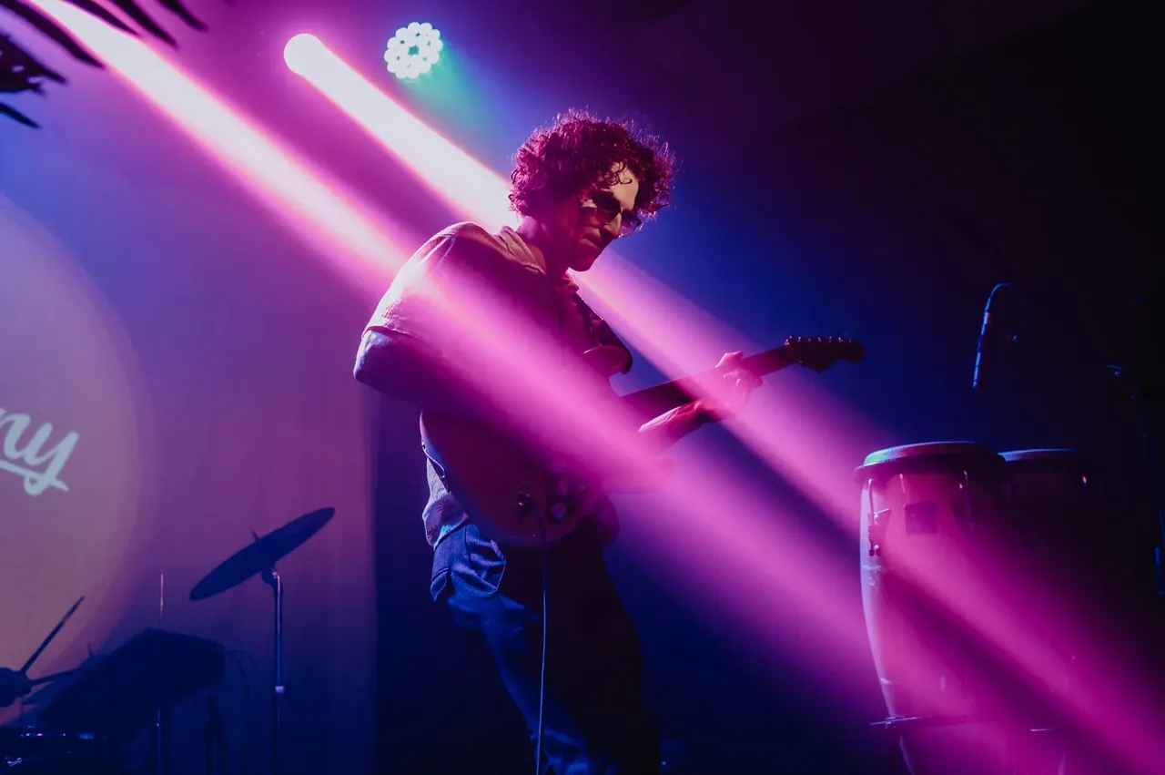 Musician performing on stage with guitar under dramatic pink and blue stage lighting, drums visible beside them.
