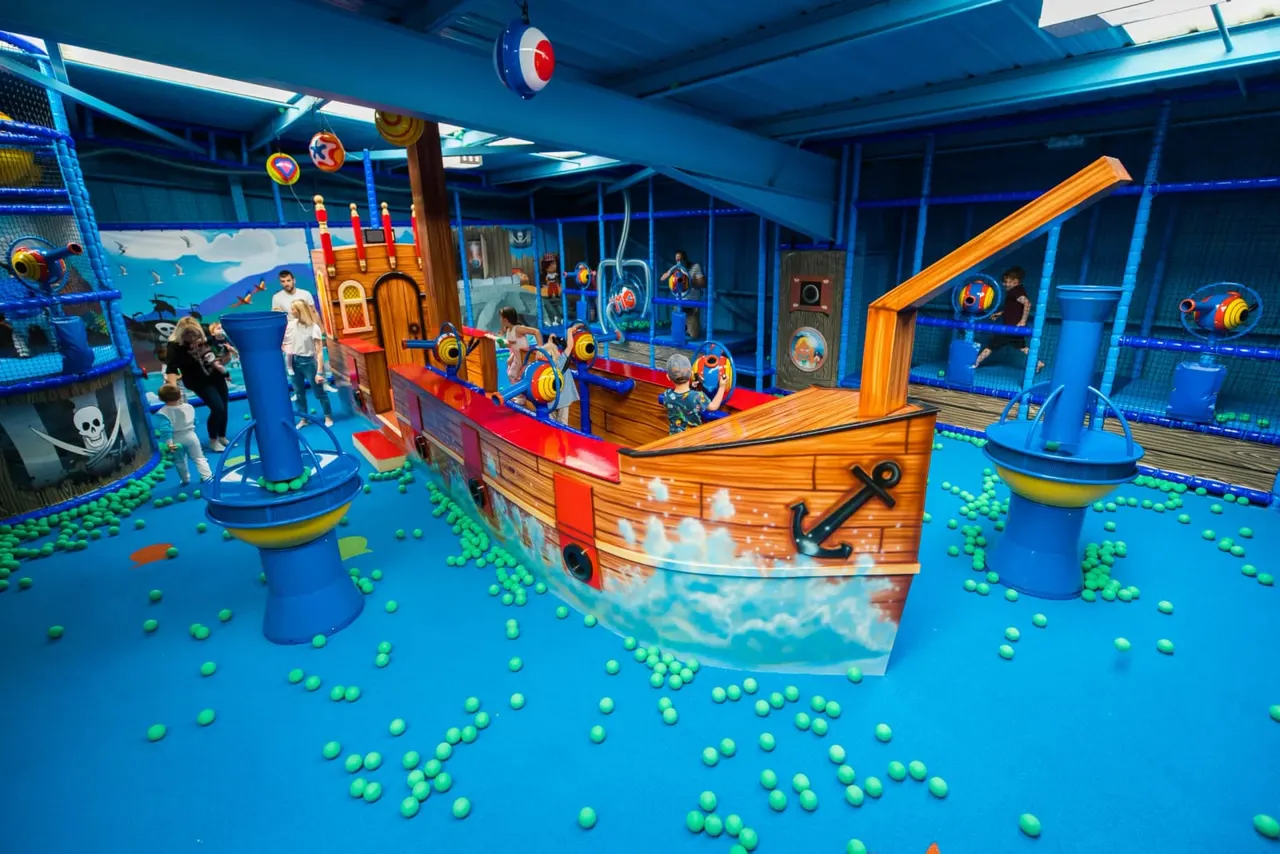 Indoor play area with children playing around a large wooden pirate ship surrounded by green balls on a blue floor.