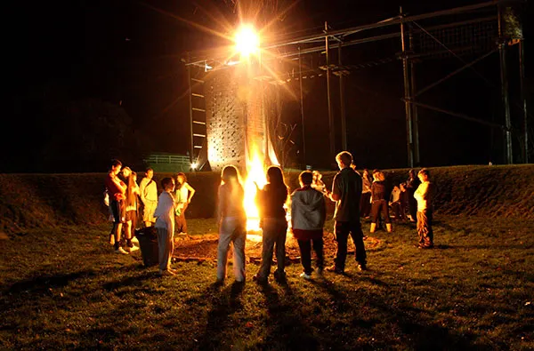 Group of teenagers gathered around a bonfire at night on sandy ground.