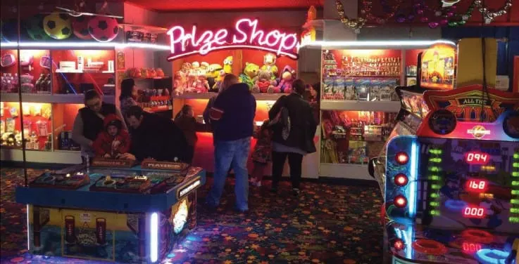 People playing arcade games and pinball machines in a brightly lit amusement arcade with a prize shop counter.