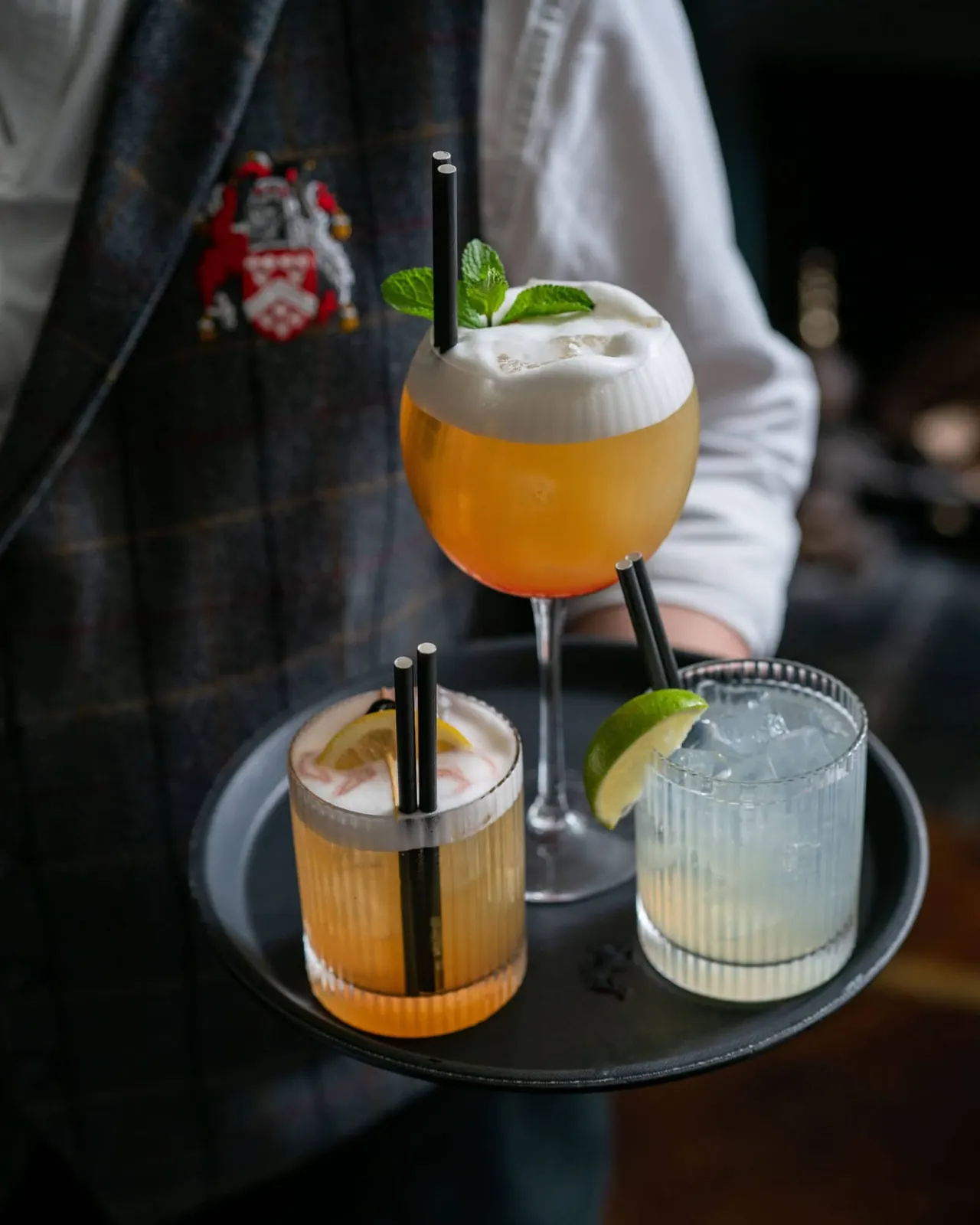 Bartender holding a tray of three colourful cocktails garnished with mint and citrus, with black straws.