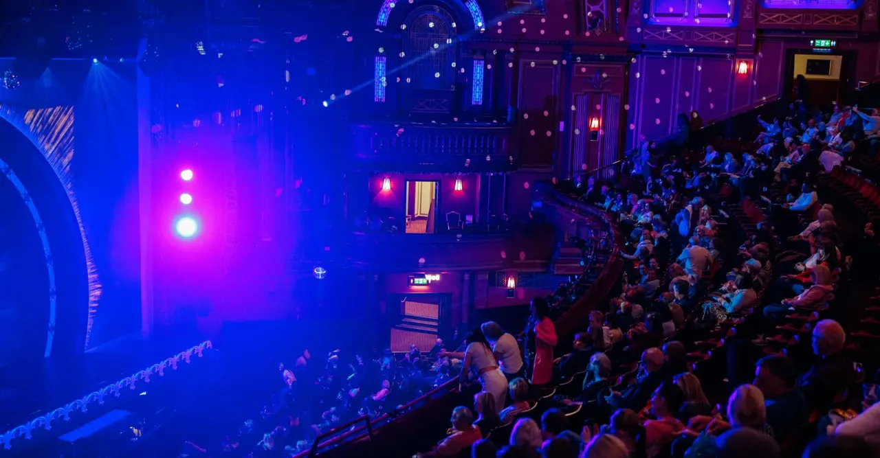 Audience seated in theatre balconies watching a stage performance lit with blue and magenta lighting effects.