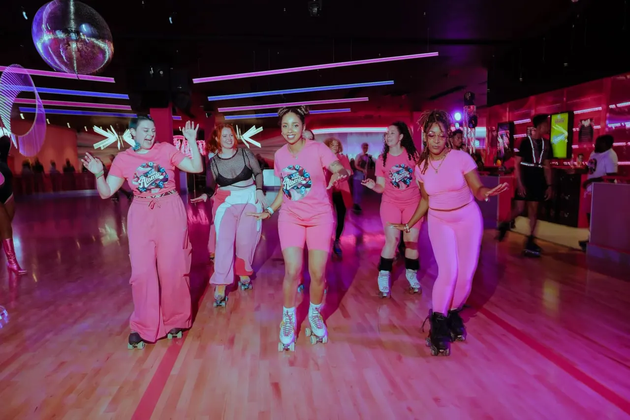 Group of people in matching pink outfits roller skating indoors under neon lighting in a vibrant venue.