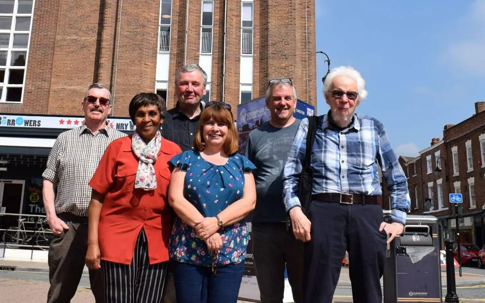 Six people of varying ages standing together on a sunny street outside a brick building.