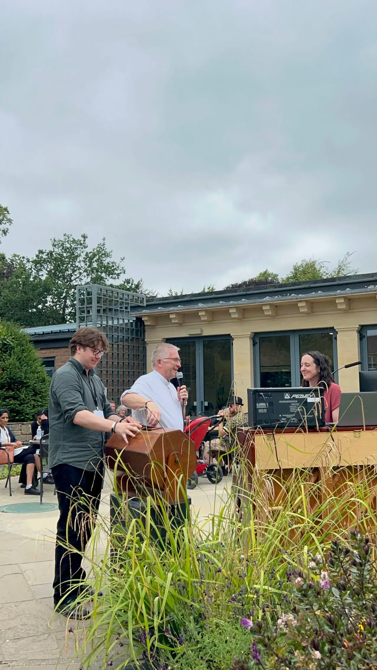 Outdoor event with three people on a paved terrace beside a modern building, surrounded by ornamental grasses and flowering plants.