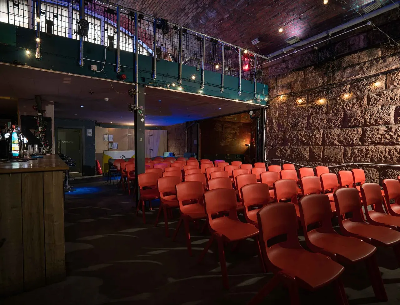 Cinema screen room with rows of red seats facing a blank screen, brick walls, bar counter on left, and mezzanine level above.