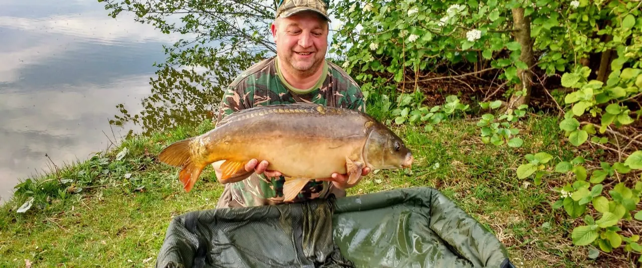 Man in camouflage clothing holding a large carp by the water's edge surrounded by green vegetation.