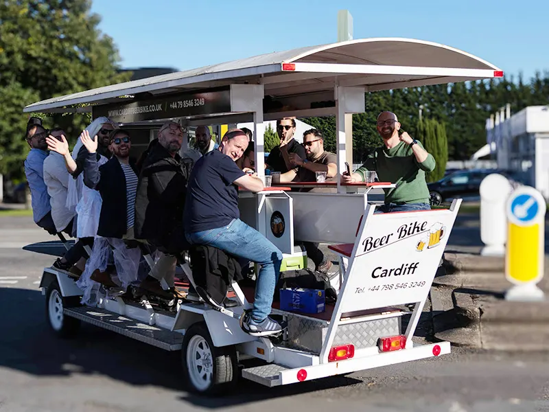 A group of people seated and standing on a multi-person pedal-powered beer bike with a bar counter, on a sunny day in a car park.
