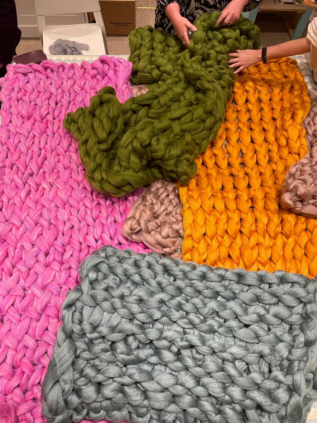Two people handling large colourful hand-knitted blankets in pink, green, orange and grey displayed on a table.
