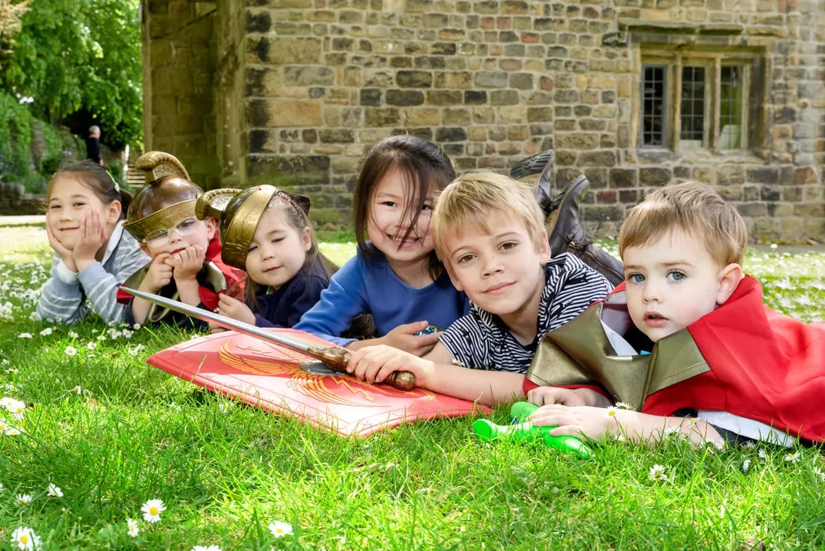 Group of children in period costumes lying on grass outside a stone building, holding a large scroll and props.