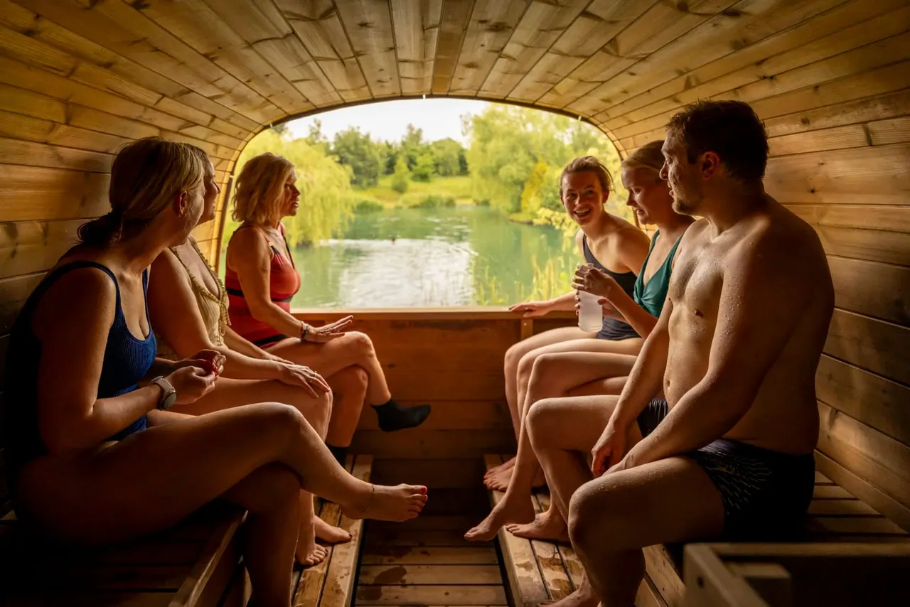 A group of five people in swimwear relaxing inside a wooden sauna hut overlooking a river and trees.