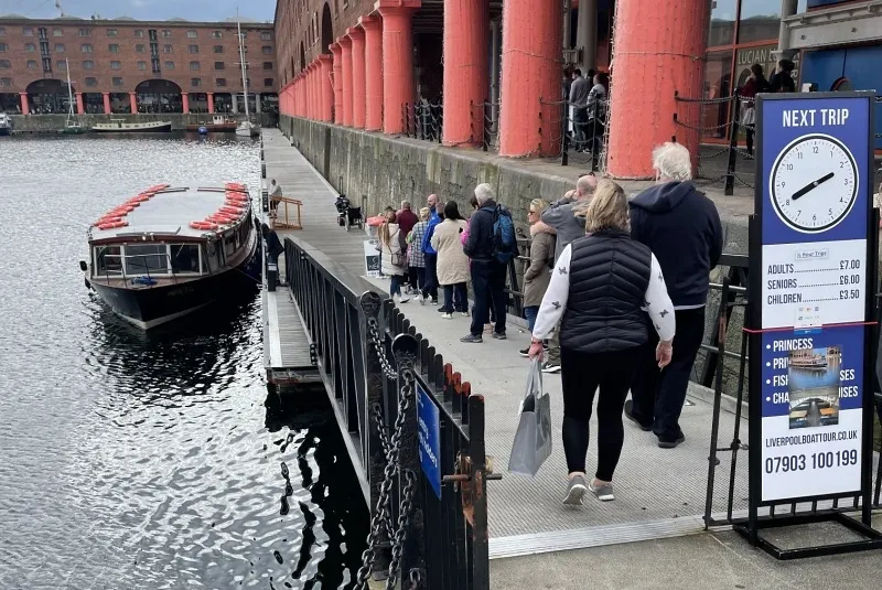 Queue of visitors boarding a tour boat at a waterfront pier with red pillars and historic buildings behind.