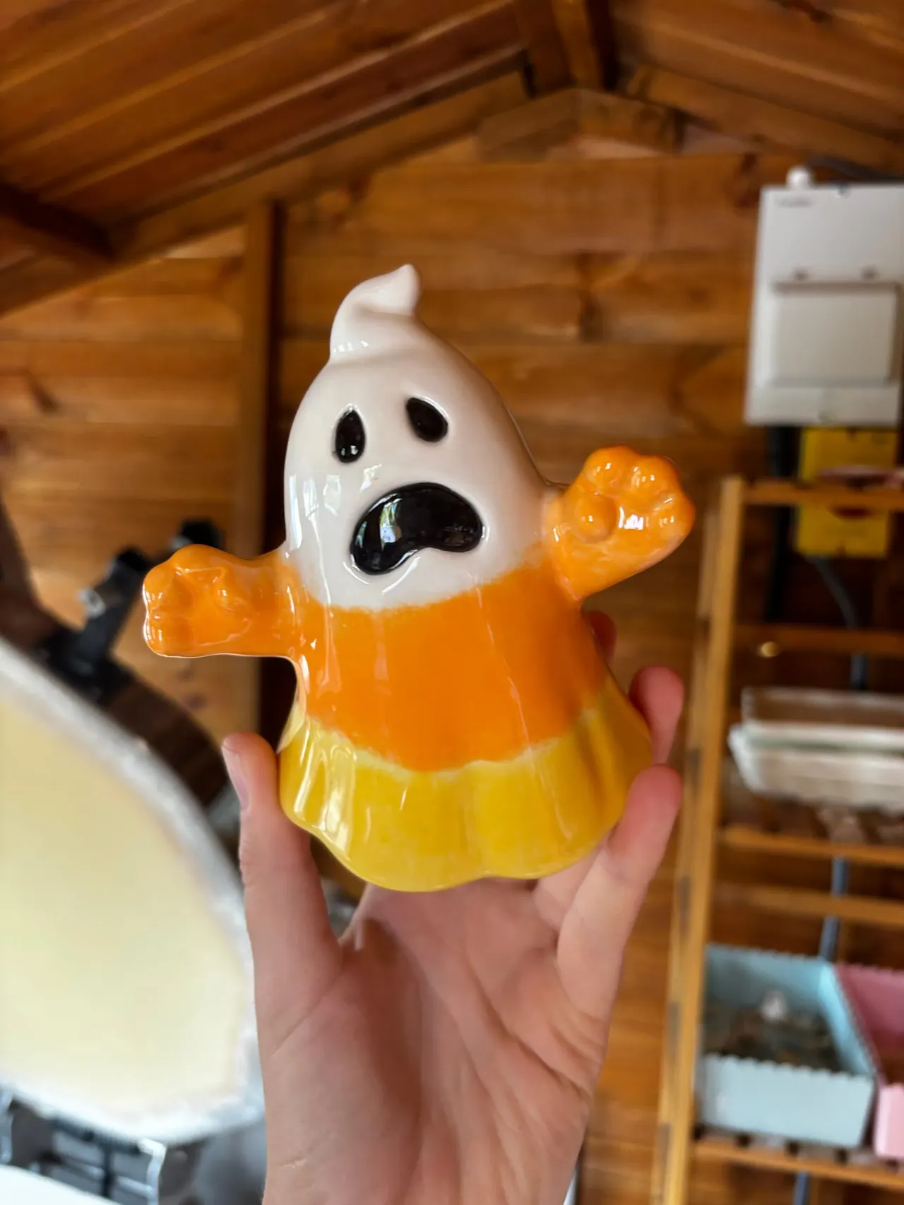 Hand holding a Halloween ghost figurine with white head, orange and yellow body, against wooden interior background.