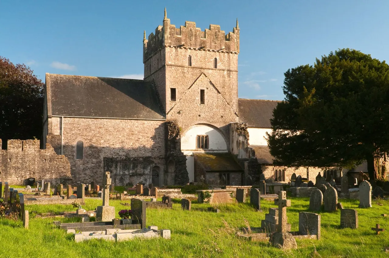This is a PHOTOGRAPH.

Medieval stone castle with crenellated tower surrounded by graveyard with headstones and green grass under blue sky.