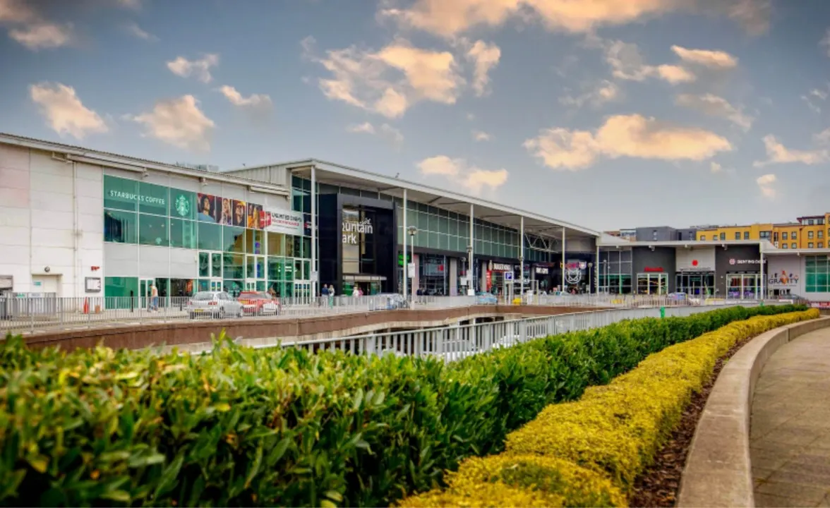 Modern shopping and leisure centre with green hedging and landscaped gardens in foreground under blue sky.