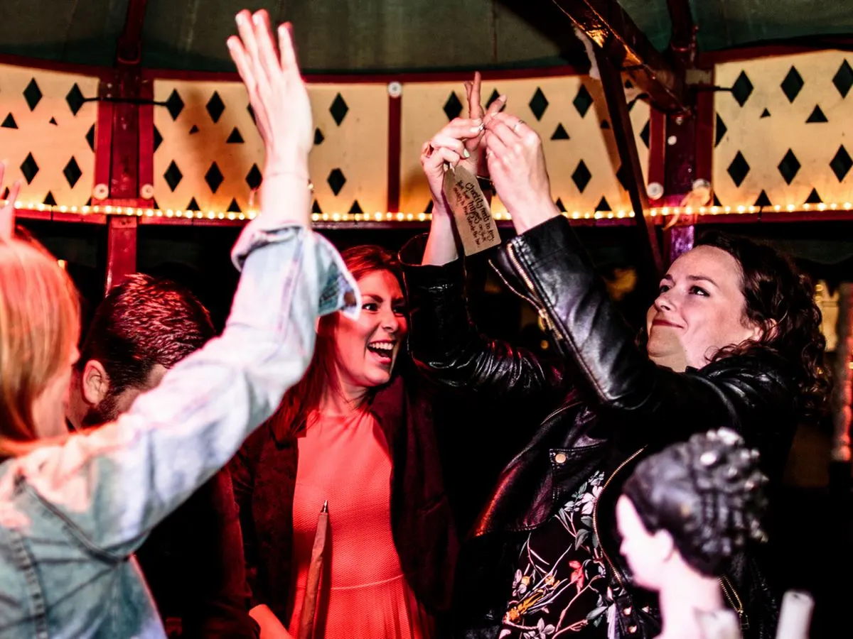 A group of people celebrate indoors under red neon lighting and a geometric patterned backdrop, with one person holding up a wristband.