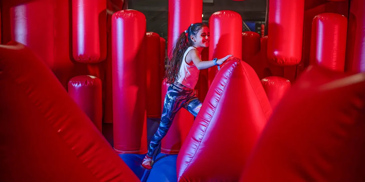 Child navigating through red inflatable obstacle course pillars in brightly lit arena.