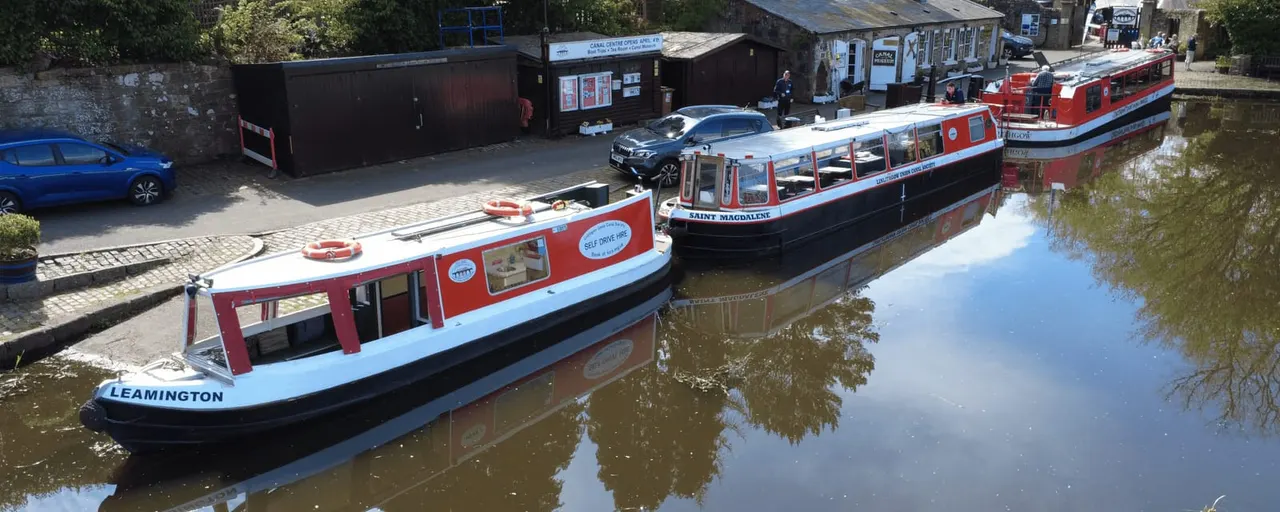 Red and white narrowboats moored on a canal with buildings and a car parked on the towpath beyond.