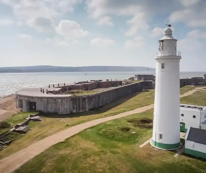 White lighthouse standing beside a coastal fort with grassy grounds overlooking a bay and distant hills.