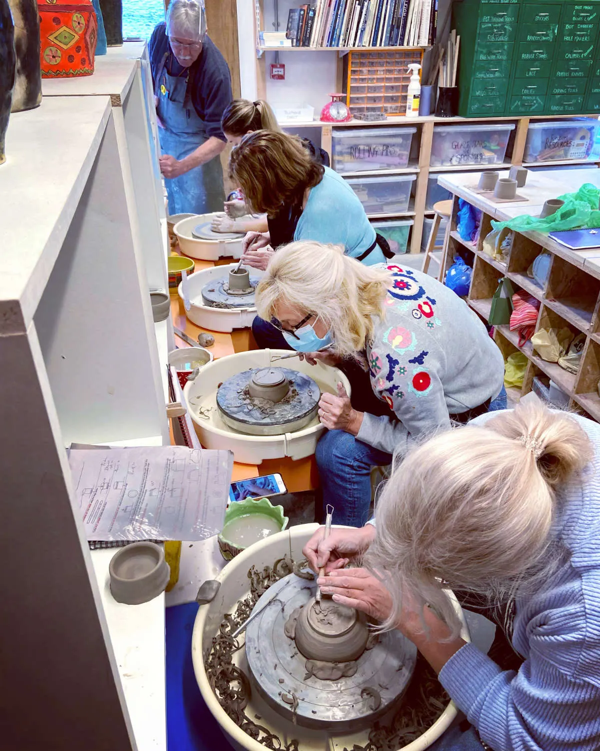 Visitors of various ages participate in a pottery wheel throwing workshop, with an instructor guiding them at clay wheels in a craft studio.