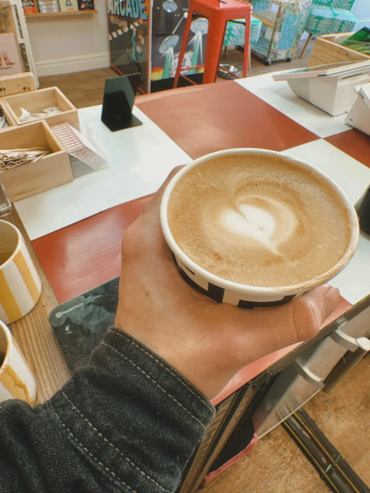 Person holding a cappuccino with latte art at a café table surrounded by board games and wooden storage boxes.
