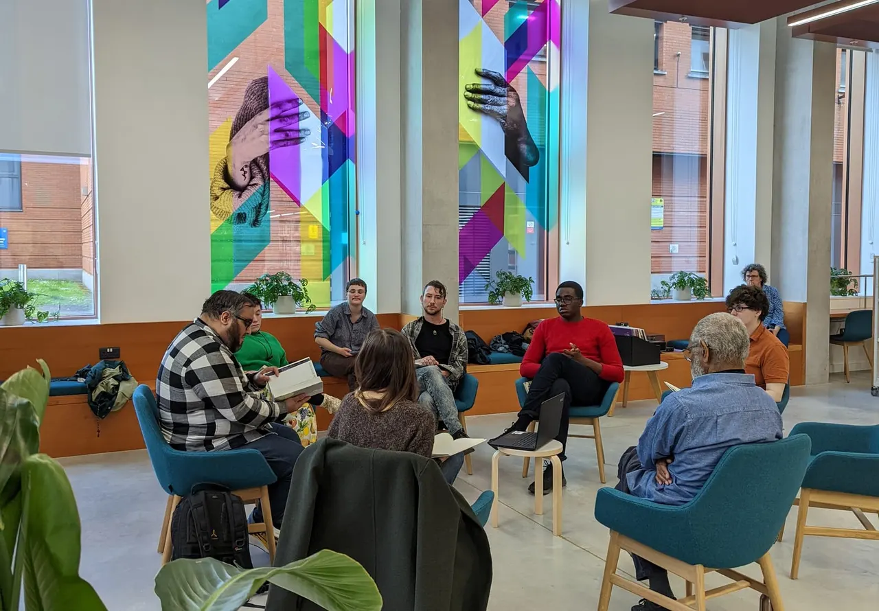 A group of people sit in a circle on teal chairs in a modern library space with colourful geometric murals on tall glass panels behind them.