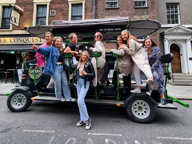 Group of people posing on a green and black pedal-powered beer bike on a city street.